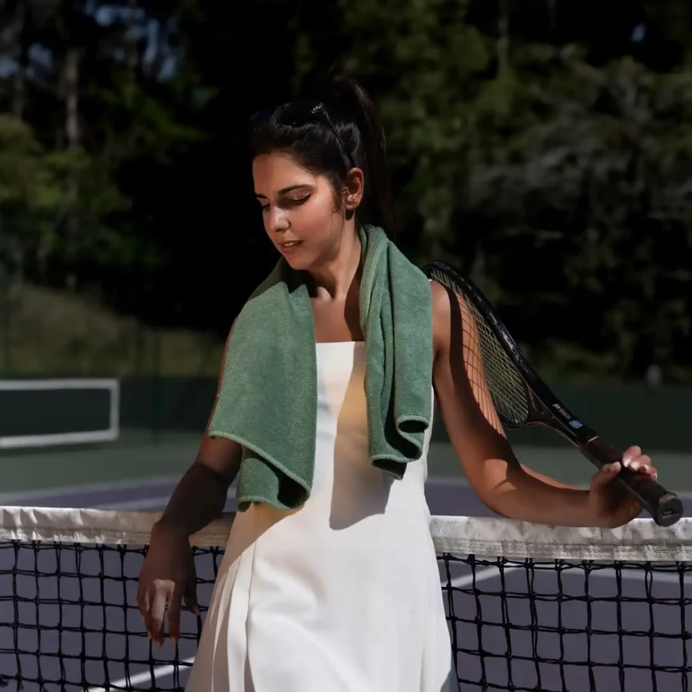 En kvinnlig tennisspelare på en tennisplan med en jadegrön Graccioza Egoist handdukar i färgen Jade hängande över axlarna som sport- och träningsinspiration.