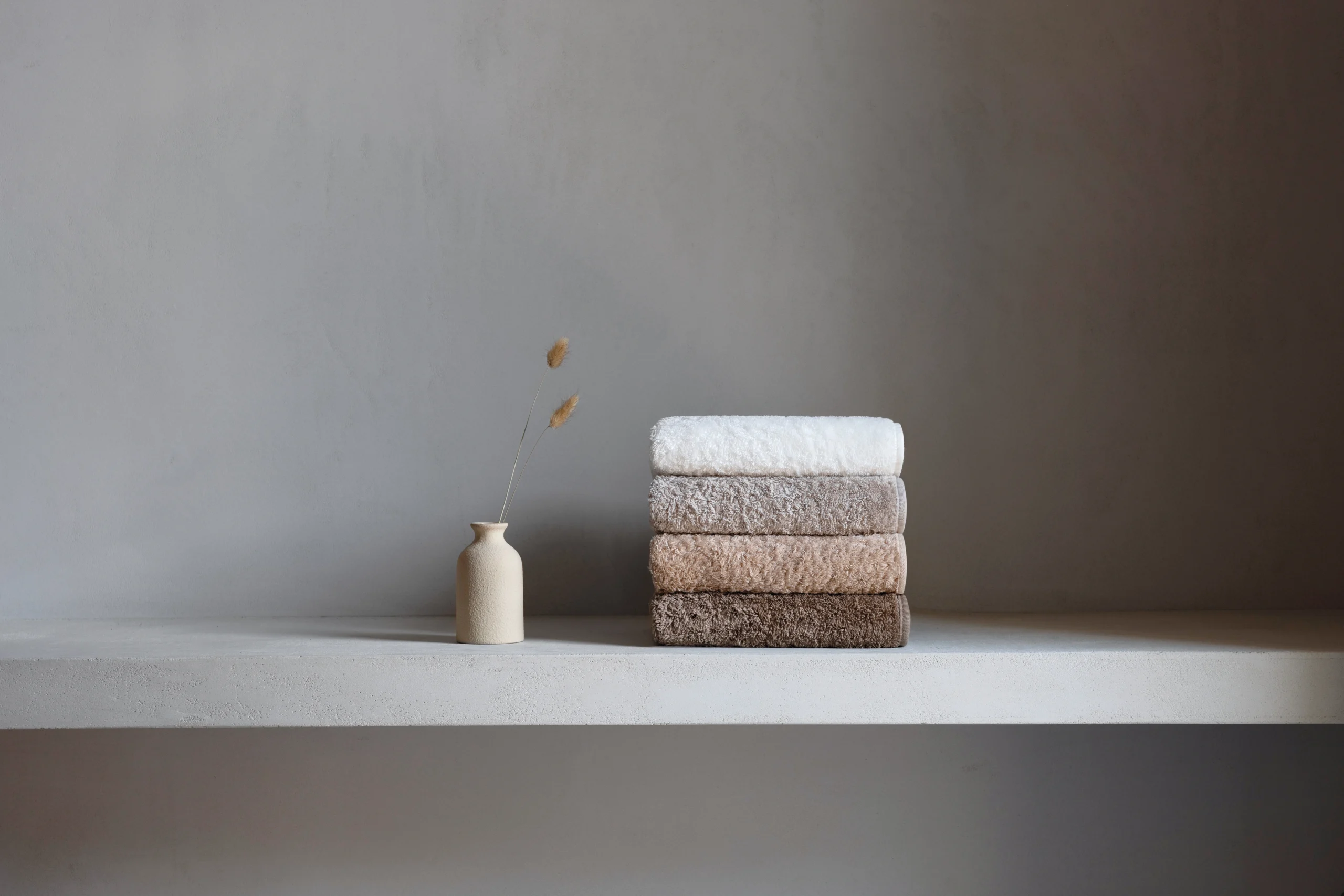Fyra exklusiva Graccioza Egoist handdukar i de skandinaviska nyanserna Snow, Fog, Linen och Stone vackert staplade på en grå hylla i ett ljust grått badrum.