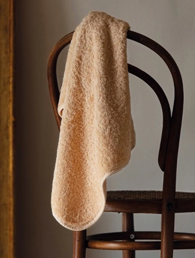 En lyxig Graccioza Egoist badhandduk i den varma, naturliga persikofärgen Toast elegant hängande över en stolsrygg i en inspirerande hemmiljö.