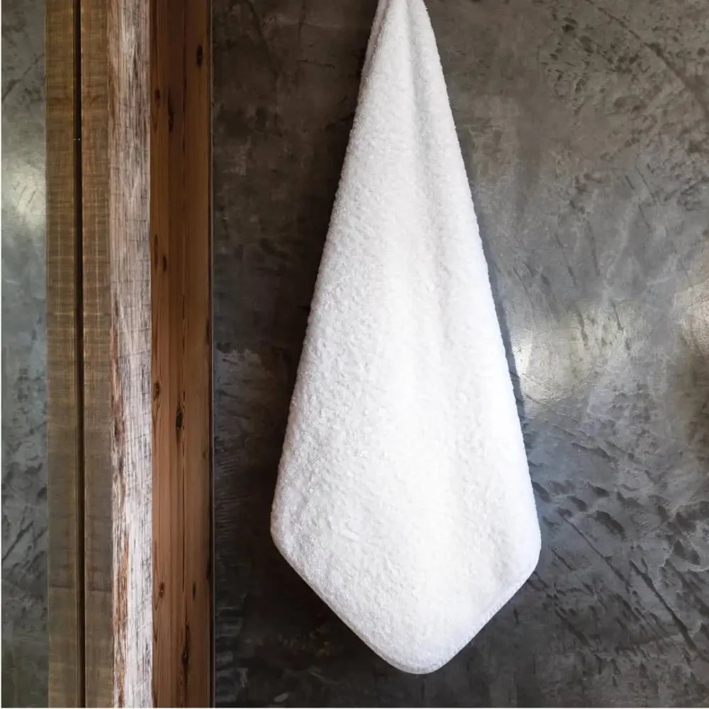 En krispigt vit Graccioza Egoist badhandduk i storleken 70x140cm upphängd på en krok i ett mörkt inrett badrum som visar en stilren visuell kontrast.